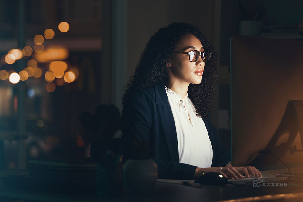 A woman in a suit is sitting at her desk, looking at her computer screen. She has brown skin, long black curly hair, and is wearing glasses. She is working late and the office is empty, except for the lights from the city outside.