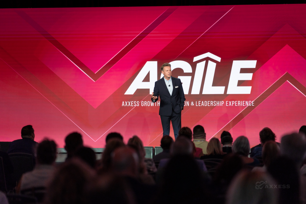 A man in a suit is standing on a stage in front of a large screen with the word "AGILE" on it. He is speaking to a large audience.