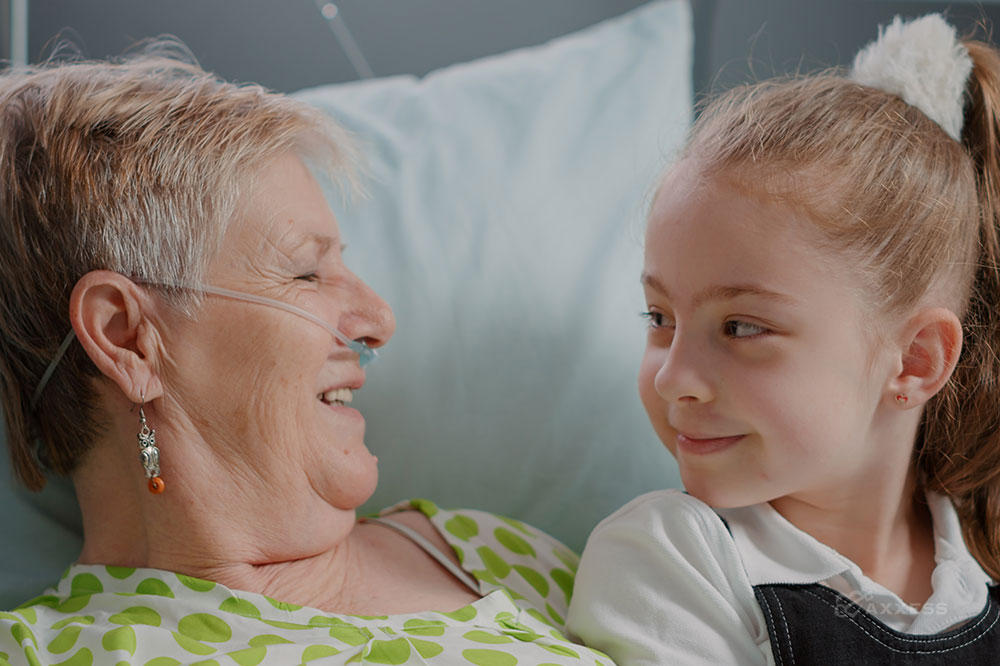 An elderly woman with oxygen tubes in her nose looks fondly at a young girl sitting on her lap