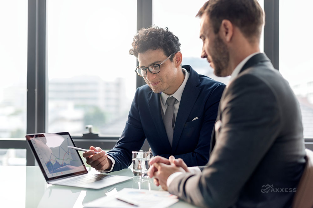 Two men in suits sit at a table and look over financial information on a computer
