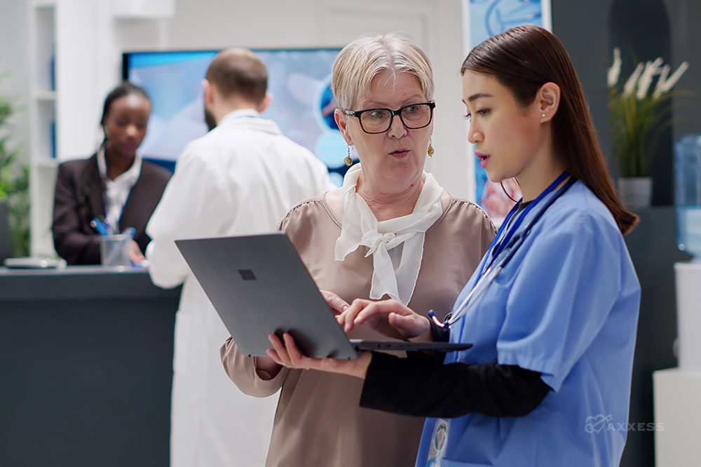 A woman in scrubs holds a laptop and speaks with another woman in professional clothing.