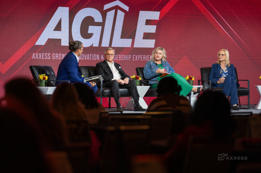 A&nbsp;panel&nbsp;of&nbsp;four&nbsp;speakers&nbsp;is&nbsp;seated&nbsp;on&nbsp;a&nbsp;stage&nbsp;in&nbsp;front&nbsp;of&nbsp;a&nbsp;large&nbsp;red&nbsp;and&nbsp;white&nbsp;sign.&nbsp;The&nbsp;speakers&nbsp;are&nbsp;arranged&nbsp;in&nbsp;a&nbsp;row&nbsp;of&nbsp;black&nbsp;chairs,&nbsp;with&nbsp;a&nbsp;man&nbsp;on&nbsp;the&nbsp;left,&nbsp;a&nbsp;man&nbsp;in&nbsp;the&nbsp;center-left,&nbsp;a&nbsp;woman&nbsp;in&nbsp;the&nbsp;center right,&nbsp;and&nbsp;a&nbsp;woman&nbsp;on&nbsp;the&nbsp;right.&nbsp;The&nbsp;audience&nbsp;is&nbsp;visible&nbsp;in&nbsp;the&nbsp;foreground,&nbsp;mostly&nbsp;blurred,&nbsp;with&nbsp;people&nbsp;seated&nbsp;in&nbsp;rows&nbsp;of&nbsp;chairs.&nbsp;The&nbsp;lighting&nbsp;is&nbsp;bright&nbsp;on&nbsp;the&nbsp;stage,&nbsp;and&nbsp;the&nbsp;overall&nbsp;tone&nbsp;of&nbsp;the&nbsp;image&nbsp;is&nbsp;professional&nbsp;and&nbsp;informative.&nbsp;The&nbsp;sign&nbsp;behind&nbsp;them&nbsp;reads&nbsp;"Axxess&nbsp;Growth&nbsp;Innovation&nbsp;&&nbsp;Leadership&nbsp;Experience".