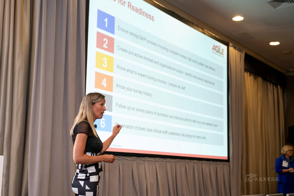 A woman presents in a conference room, standing beside a projection screen displaying a slide titled Tips for Readiness. The slide lists six color-coded items related to survey readiness, including operational processes, preparation calendars, survey history, and staff alignment. A laptop sits on a podium, and another person stands in the background. Rows of chairs face the presenter.