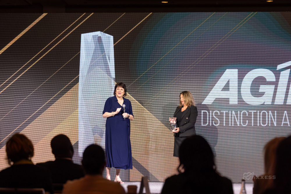 Award ceremony at the 2025 Axxess AGILE conference in Dallas, Texas, featuring the presentation of the AGILE Distinction Award on stage. Two individuals stand at the center, one speaking into a microphone, the other holding the trophy, while a digital display and audience are visible in the background.