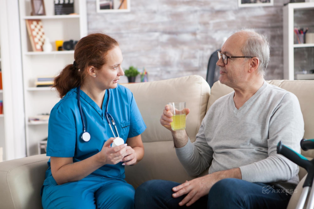 A healthcare professional in blue scrubs sits on a couch beside an older man in a home setting, holding a medication bottle while the man listens and holds a glass of yellow liquid.