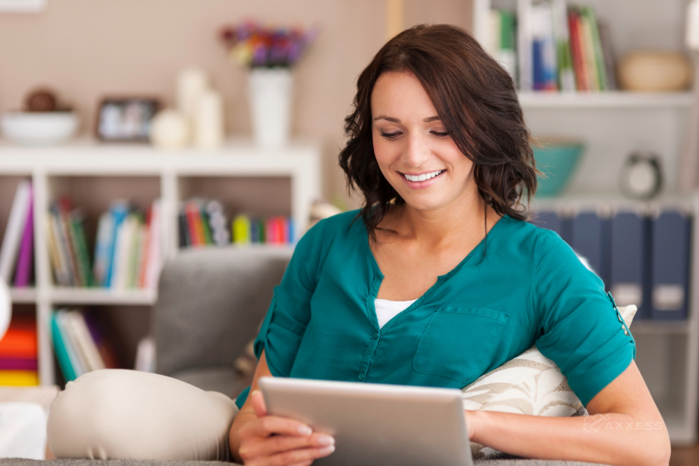 Person sitting on a couch in a cozy living room, holding a tablet and looking at the screen, with a bookshelf and decorative items in the background.