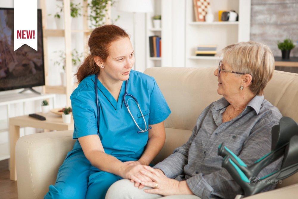 A healthcare professional in blue scrubs sits on a couch beside an older adult, gently holding their hand and speaking with them in a warm, supportive manner. They are in a cozy living room with a television, bookshelves, and a potted plant in the background, conveying a calm, home‑based care setting.