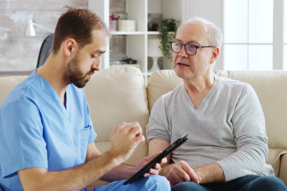 Home healthcare worker in blue scrubs sits beside an older adult on a couch in a living room, using a tablet while discussing care. A walker is partially visible next to the couch, and shelving and home furnishings appear in the background.
